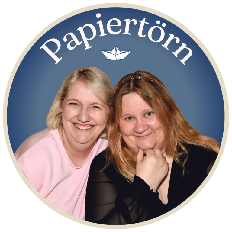 Anja und Petra sitzen lächelnd dicht beieinander vor einem blauen, runden Hintergrund, über dem das Wort „Papiertörn“ und die Abbildung eines Papierboots zu sehen sind, und feiern den bevorstehenden Papiertörn-Basteltag 2026.