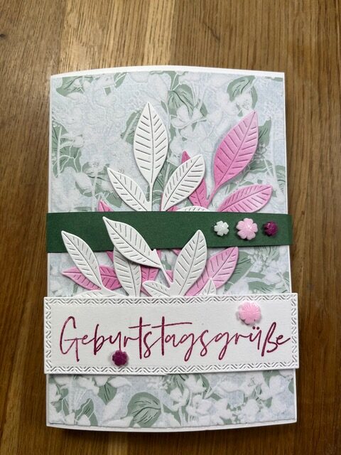 Eine handgemachte Geburtstagskarte, inspiriert von Mach, vom 26. Mai, mit weißen und rosa Papierblättern, kleinen Blumenverzierungen und dem Schriftzug „Geburtstagsgrüße“ in roter Schreibschrift auf der Vorderseite.