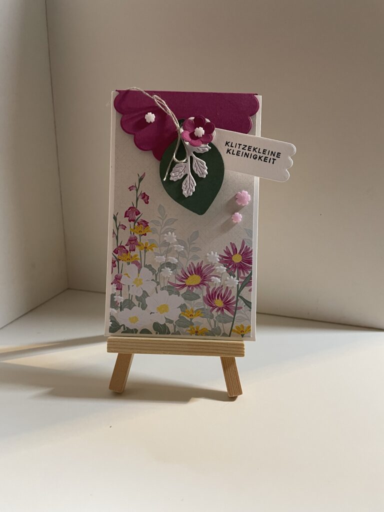 Eine handgefertigte Grußkarte mit Blumenmotiven und Verzierungen aus rosa und grünem Papier, angefertigt für den 26. Mai, mit einem kleinen Anhänger mit der Aufschrift „KLITZEKLEINE KLEINIGKEIT“, präsentiert auf einer Miniatur-Holzstaffelei.