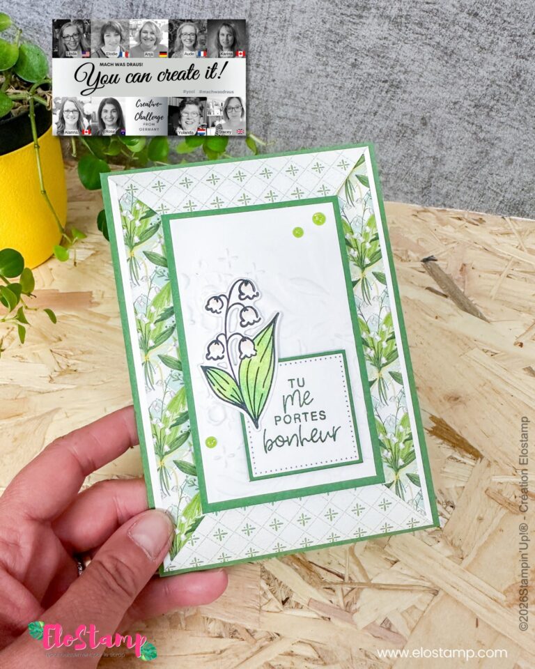 Eine Hand hält eine Grußkarte mit Blumenmotiv und der Aufschrift „Tu me portes bonheur“, die auf einer Holzunterlage neben einem gelben Blumentopf mit einer Pflanze zu sehen ist. Oben ist ein Banner mit einem Gruppenfoto und der Aufschrift „Mach was draus 26. Juni“ zu sehen.