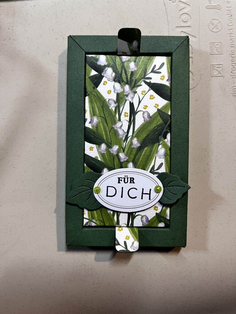 Eine grüne, rechteckige Karte von Mach aus dem 26. Juni zeigt ein Blumenmotiv und den Schriftzug „FÜR DICH“ auf einem weißen, ovalen Etikett, das mit zwei grünen Blattformen verziert ist.