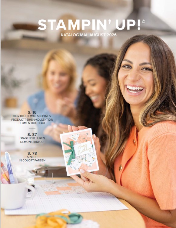 Drei Frauen sitzen an einem Tisch und basteln handgemachte Karten. Die Frau im Vordergrund lächelt in die Kamera und hält eine verzierte Karte in der Hand – vielleicht inspiriert durch den „Stampin’ Up!-Katalog 2026“. Die Atmosphäre ist fröhlich und kreativ.