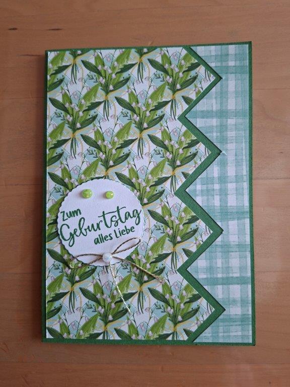 Eine handgefertigte Geburtstagskarte mit grünen Blatt- und Karomuster, inspiriert von „Mach war draus“ vom 26. Juni, mit einem runden Aufkleber, auf dem auf Deutsch „Zum Geburtstag alles Liebe“ steht.