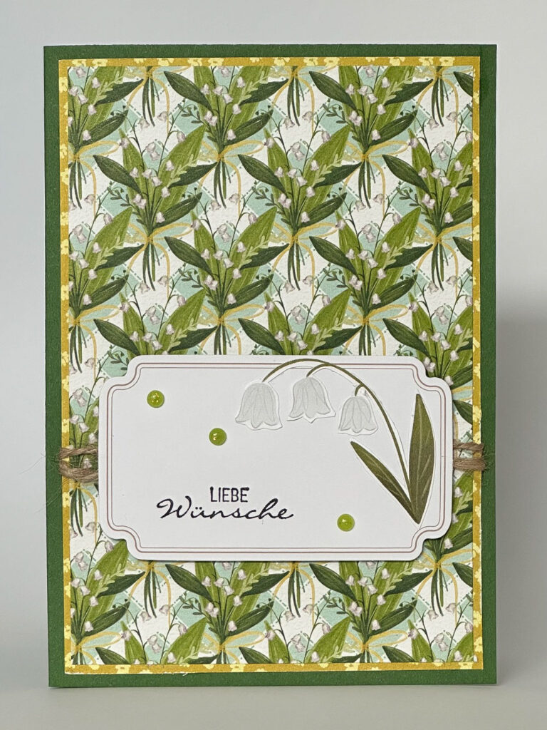 Eine Grußkarte mit grünem Blumenmuster, versehen mit einem weißen Etikett mit Blumenillustrationen, grünen Punkten und dem deutschen Text „Liebe Wünsche“ – inspiriert von „Mach was draus, 26. Juni“.