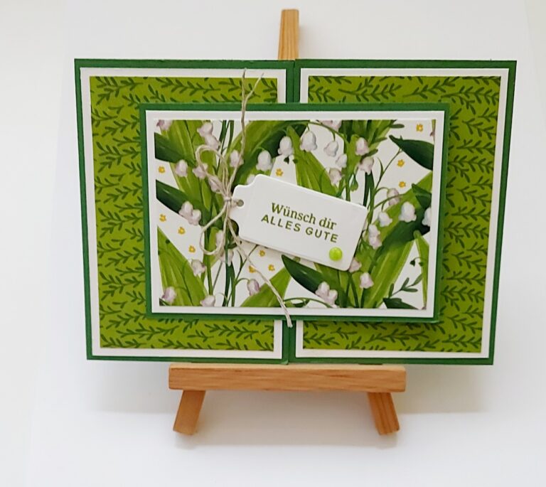 Auf einer kleinen Holzstaffelei steht eine handgemachte Karte mit grünen Blattmustern und weißen Blumen. Auf einem Anhänger an der Karte steht „Wünsch dir ALLES GUTE“. Perfekt als kreatives Projekt für „Mach was draus“ am 26. Juni.