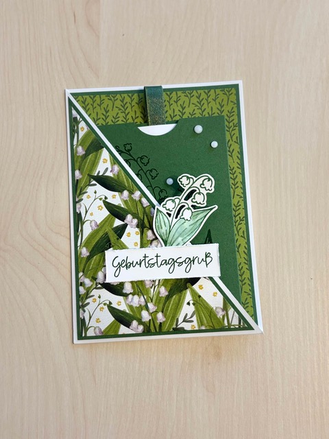 Eine handgefertigte grüne Geburtstagskarte, die für den 26. Juni entworfen wurde, zeigt Maiglöckchen-Motive, einen „Geburtstagsgruß“-Schriftzug und dekorative Verzierungen auf einer Holzunterlage.