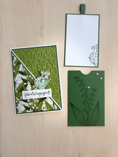 Eine handgefertigte Geburtstagskarte und dazu passende grüne Papiereinlagen, allesamt mit Maiglöckchen-Motiven verziert, präsentiert auf einer hellen Holzunterlage – perfekt als Inspiration für Ihren „Mach was draus“-Beitrag am 26. Juni.