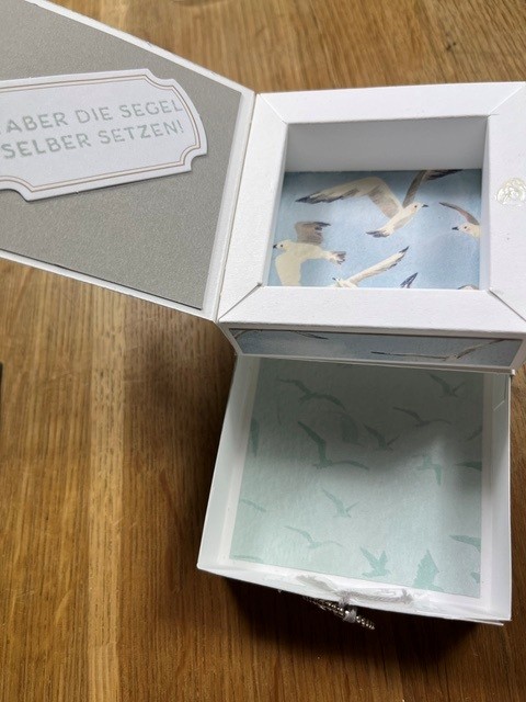 Offene weiße Geschenkbox mit Möwenmotiven im Inneren; auf dem Deckel steht auf grauem Hintergrund der deutsche Text „ABER DIE SEGEL SELBER SETZEN!“. Die Box, die Teil der Kollektion „Mach was draus – 26. Juli“ ist, steht auf einer Holzunterlage.