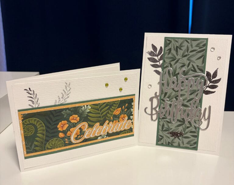 Zwei handgefertigte Grußkarten mit grünen Blumenmotiven – perfekt für „Mach was draus, 26. August“. Auf der einen Karte steht „Celebrate“ und auf der anderen „Happy Birthday“ in Metallic-Schrift, beide mit dekorativen Blättern verziert.