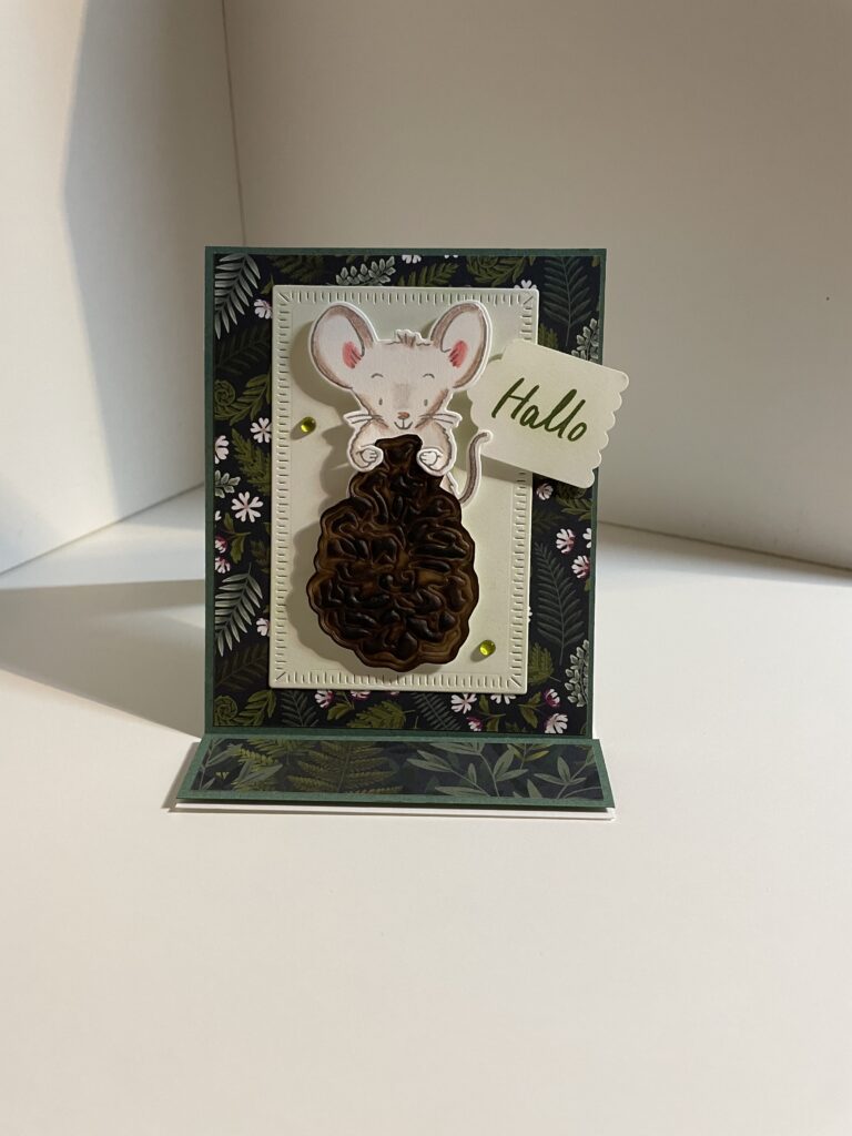 Eine Grußkarte mit der Zeichnung einer Maus, die einen Tannenzapfen hält, und einem Anhänger mit der Aufschrift „Hallo“ vor einem Blumenhintergrund – perfekt, um ein wenig Kreativität im Sinne von „Mach was draus“ am 26. August zu verbreiten.