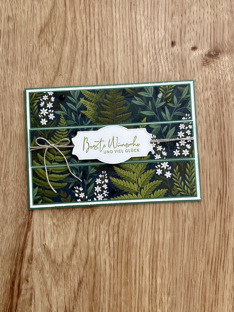 Eine Grußkarte mit einem grünen botanischen Motiv, einer Schnur-Schleife und einem Etikett mit der Aufschrift „Beste Wünsche und viel Glück“ auf Deutsch ist für „Mach was draus“ am 26. August wunderschön auf einer Holzunterlage präsentiert.