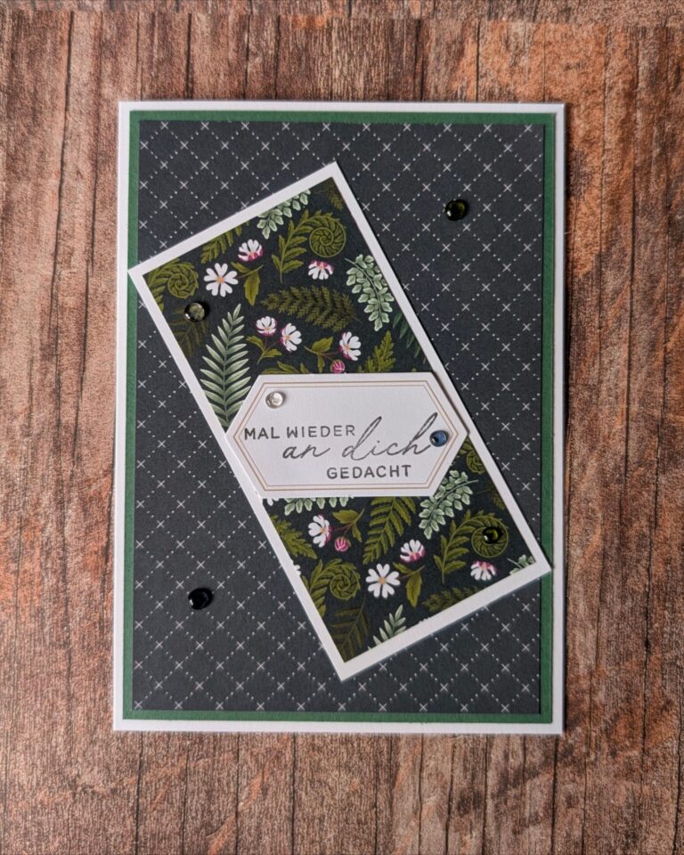Grußkarte mit botanischem Muster und dem deutschen Text „Mal wieder an dich gedacht“ auf einem weißen Etikett, das auf einer Holzunterlage angebracht ist – die perfekte Inspiration für dein nächstes kreatives Projekt oder die „Mach was draus“-Challenge am 26. August.