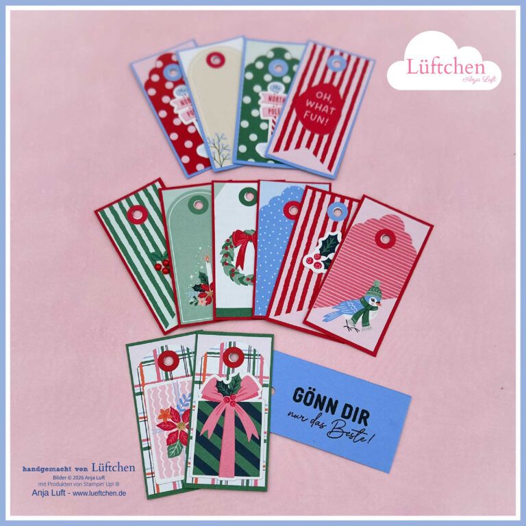 Zwölf farbenfrohe Geschenkanhänger mit festlichen Motiven und Streifen sind halbkreisförmig angeordnet, inspiriert vom „Stempelnd durchs neue Herbst-Winter-Katalog 26“, wobei unten rechts eine blaue Karte mit der Aufschrift „GÖN DIR nur das Beste!“ zu sehen ist.
