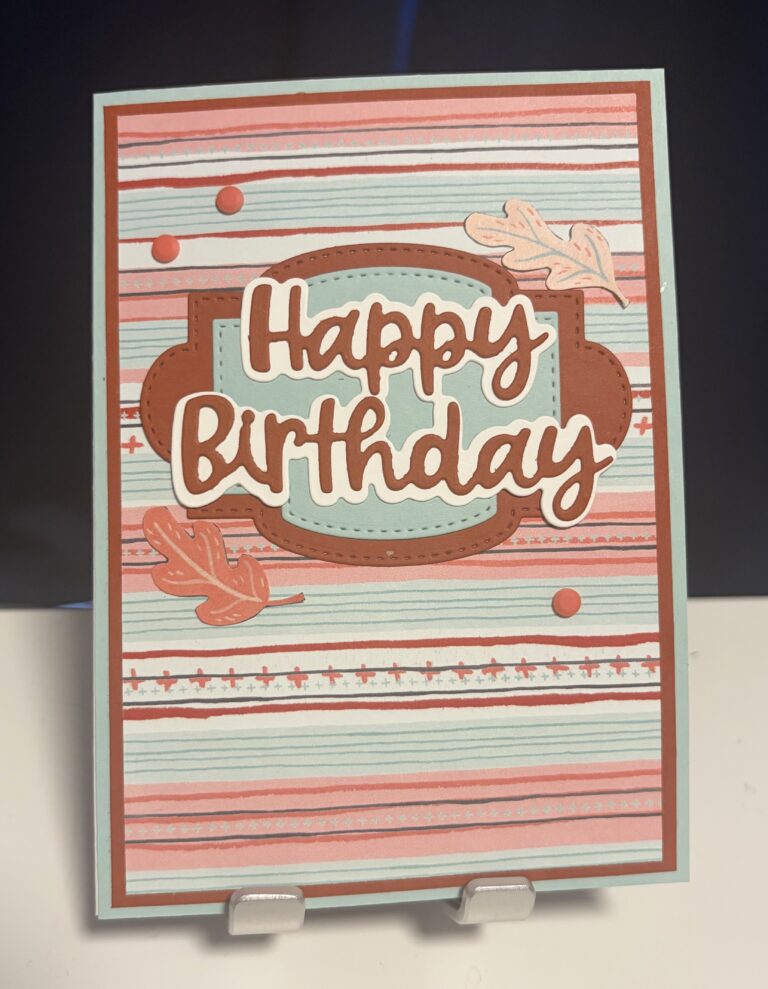 Eine handgefertigte Geburtstagskarte mit der Aufschrift „Happy Birthday“, verziert mit Blättern, Linien und Punkten in Rot-, Blau- und Brauntönen – perfekt für den 26. September. Präsentiert auf einem weißen Ständer.