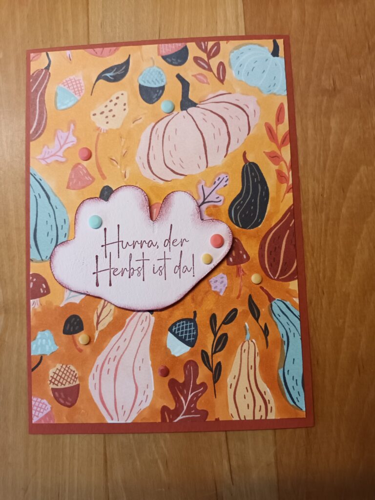 Eine Grußkarte mit farbenfrohen Illustrationen zum Thema Herbst und einem blattförmigen Ausschnitt mit der Aufschrift „Hurra, der Herbst ist da!“ – perfekt für „Mach was draus“ am 26. September – auf einer Holzunterlage.