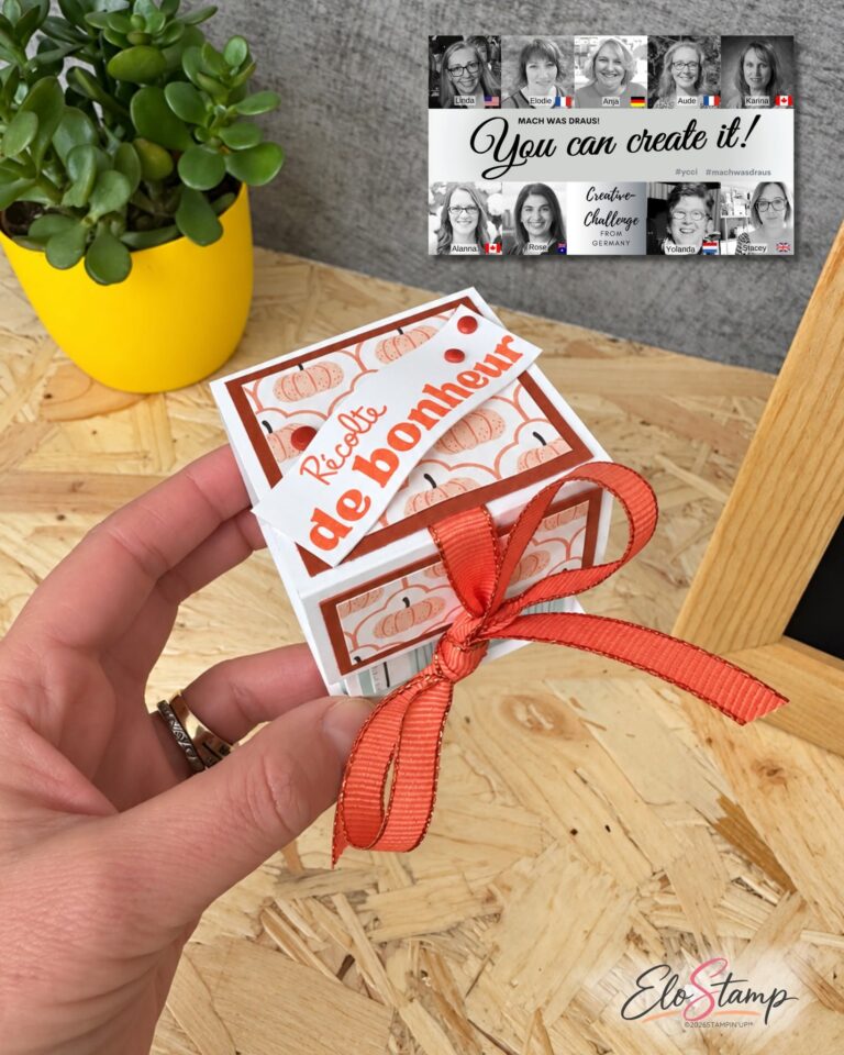 Eine Hand hält eine kleine, verzierte Geschenkbox mit einer roten Schleife; im Hintergrund sind eine Pflanze und ein Bastel-Flyer mit der Aufschrift „Mach was draus – 26. September“ zu sehen.