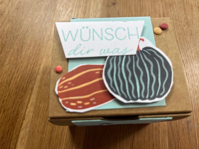 Eine kleine braune Geschenkbox, verziert mit Papierausschnitten von zwei Kürbissen und einer „Wünsch dir was“-Karte, steht auf einer Holzunterlage für „Mach was draus“ am 26. September.